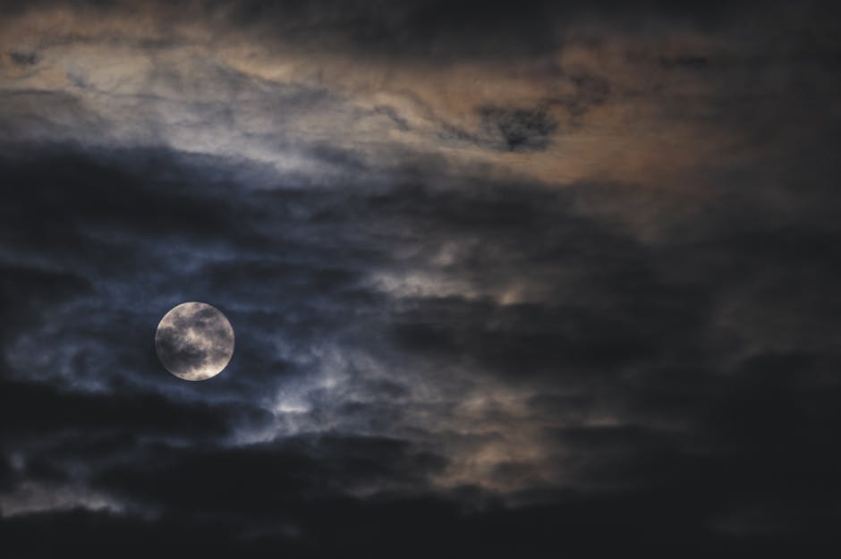 A dark, moody night sky capturing the full moon surrounded by dramatic clouds.