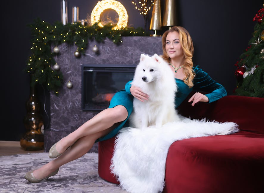 Stylish woman in a teal dress relaxes with a Samoyed dog by a festive, decorated fireplace indoors.