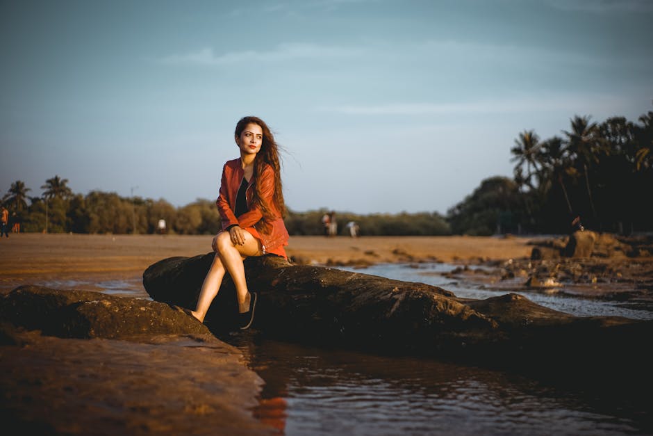 A stylish woman in outerwear sitting on rocks by the beach during sunset, creating a serene and fashionable scene.