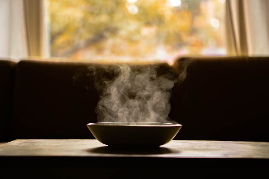 A steaming bowl of soup on a wooden table indoors with soft autumn light through a window.