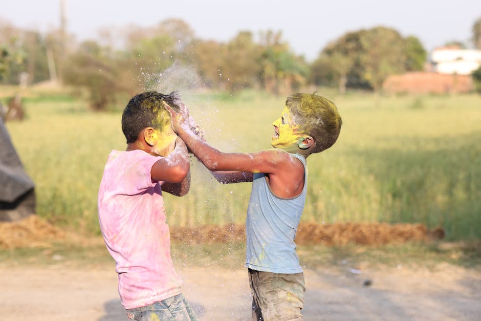 Two children playfully throw vibrant colored powders at each other in a sunny outdoor setting.