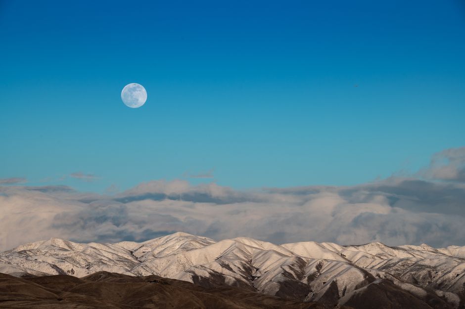 Capture of snow-covered mountains under a blue sky with the moon in view, showcasing nature's serene beauty.