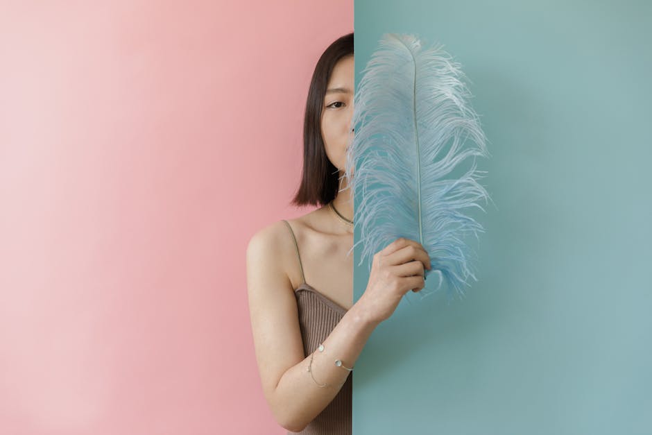 Elegant studio portrait of an Asian woman partially hidden behind a feather against pink and blue background.