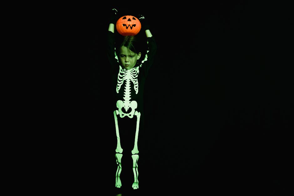 A child in a glowing skeleton costume holds a jack-o'-lantern against a black background for Halloween.