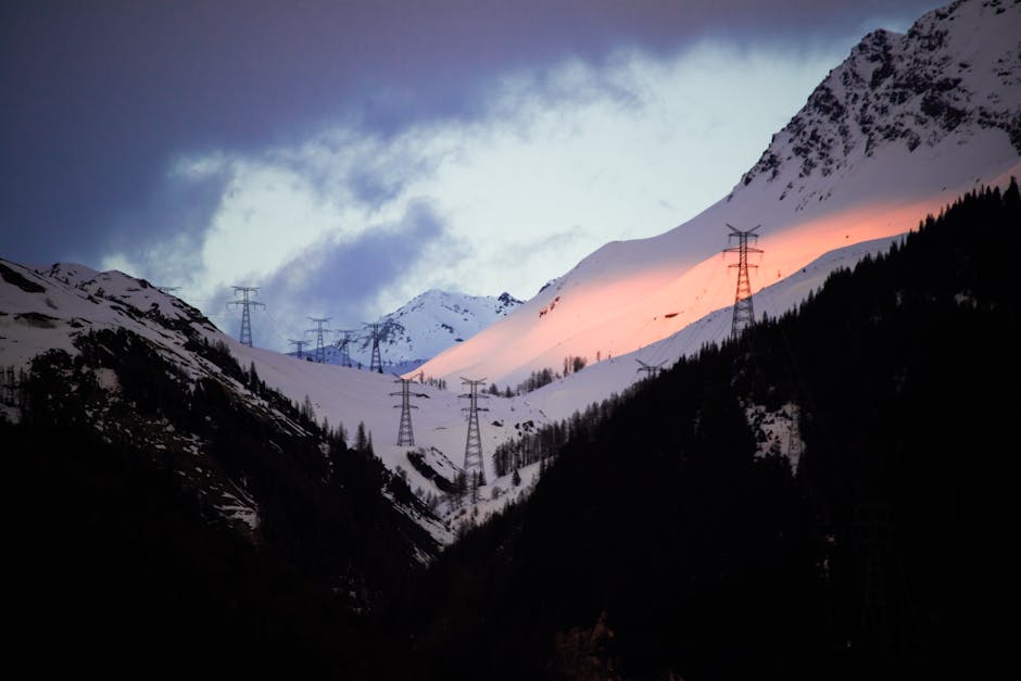 A dramatic mountain landscape at sunset with snow-covered peaks and electrical towers.