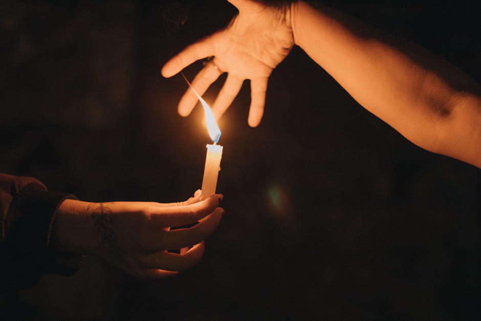 Close-up of hands sharing a lit candle in a dark setting, symbolizing connection and warmth.