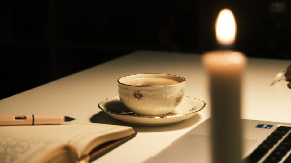 Warm candlelight illuminating a cup of tea, open notebook, and pen on a desk.