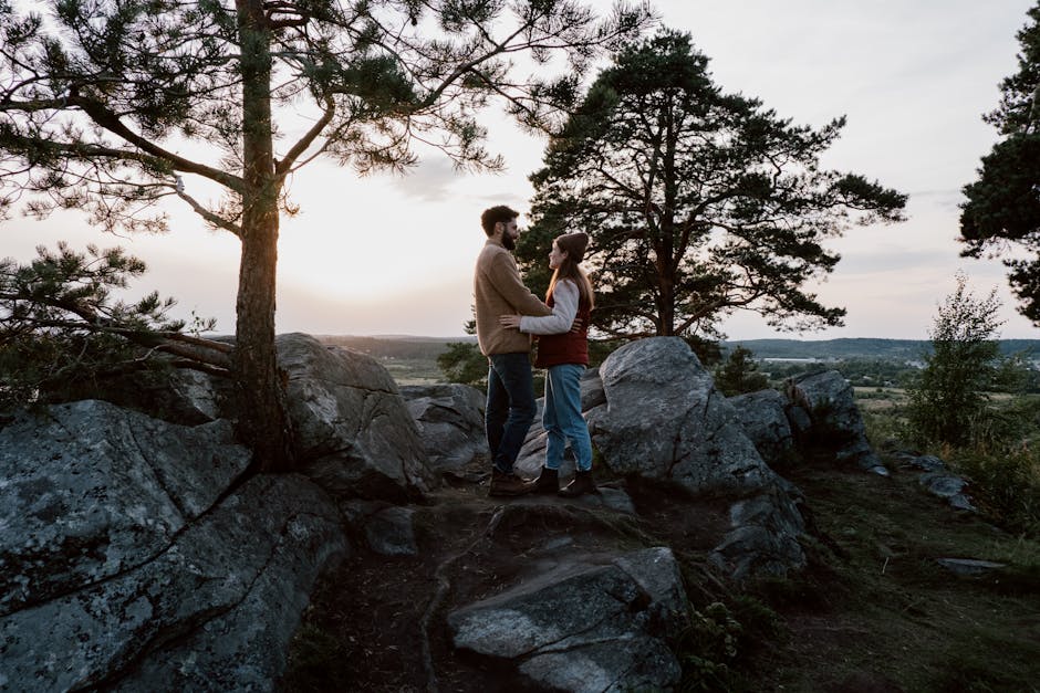 A couple embracing on a rocky hilltop surrounded by trees with a scenic sunset view.