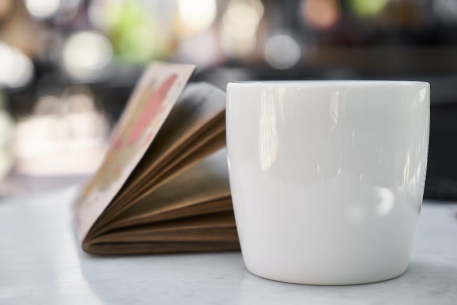 A white mug of coffee beside an open book on a marble table, indoors.