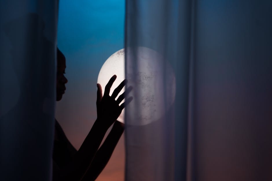 Artistic silhouette of a person holding a glowing orb behind sheer curtains at twilight.