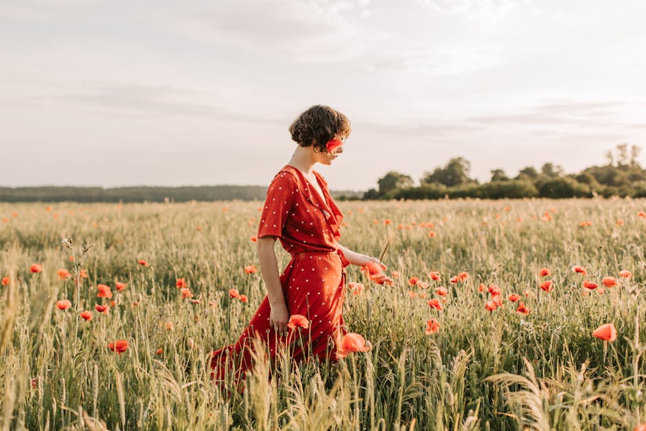 A woman in a red dress with short hair walks through a vibrant field of poppies on a sunny day.