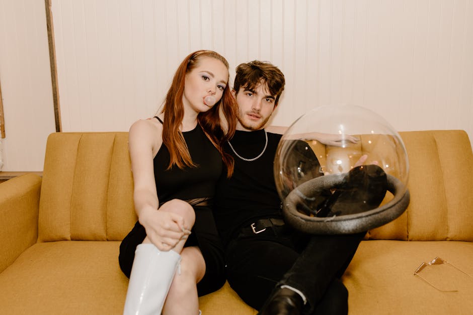 A stylish couple in futuristic attire sitting on a sofa indoors, with a space helmet.