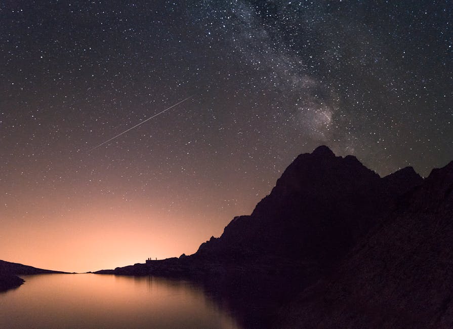 A captivating view of the Milky Way and shooting star above a silhouetted mountain reflecting over a calm lake.