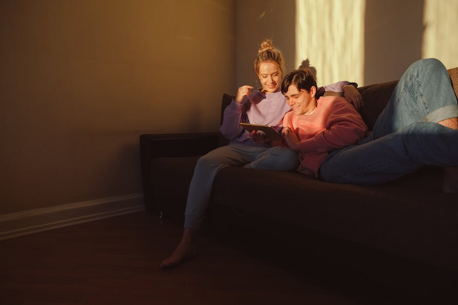 A couple sits on a sofa using a digital tablet in warm evening light, enjoying a quiet moment together.