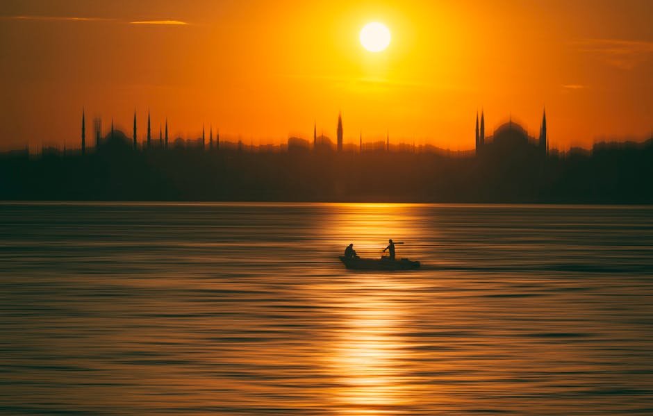 Silhouette of a fisherman in a boat against an orange sunset over a calm sea with city skyline.