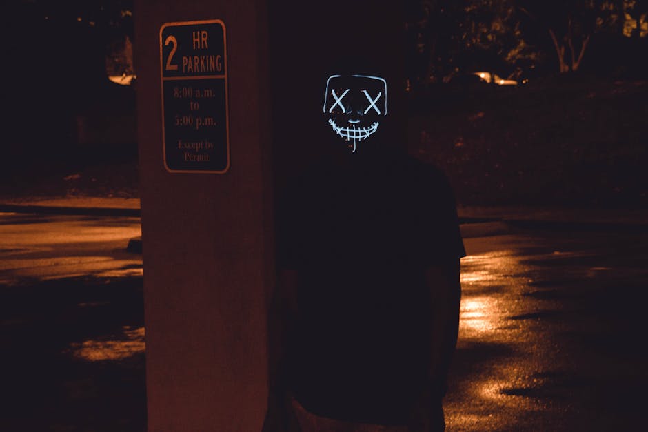 A masked person stands eerily illuminated in a parking lot, creating a spooky night scene.