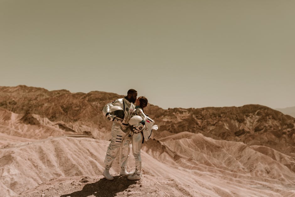 A couple in astronaut suits shares a romantic moment atop a rugged mountain landscape.