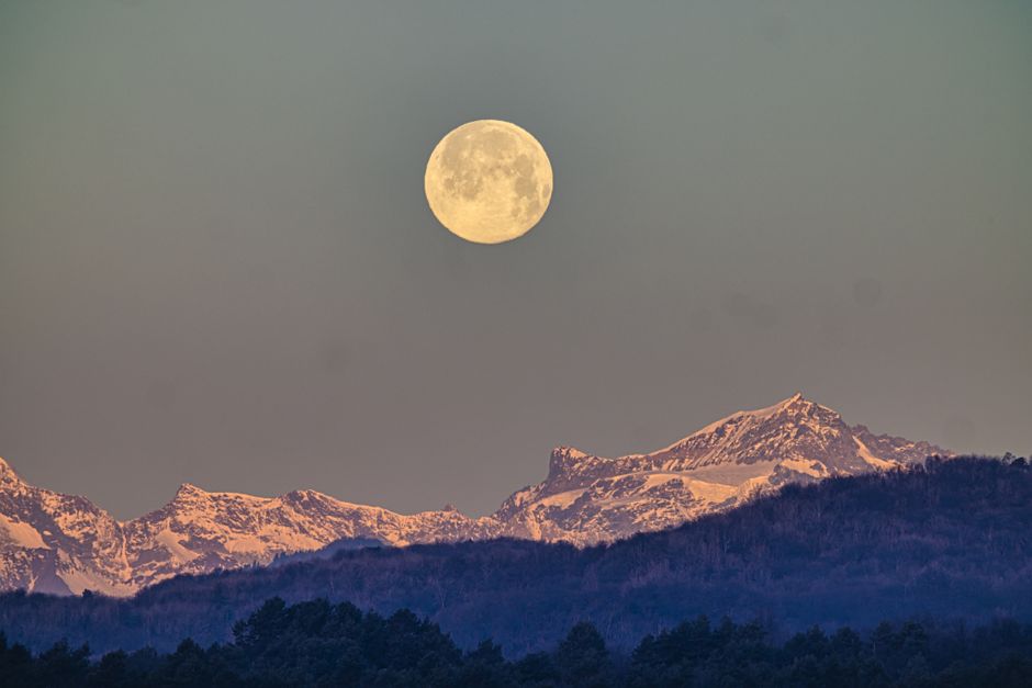 A breathtaking view of a full moon rising above snow-covered mountains during twilight.